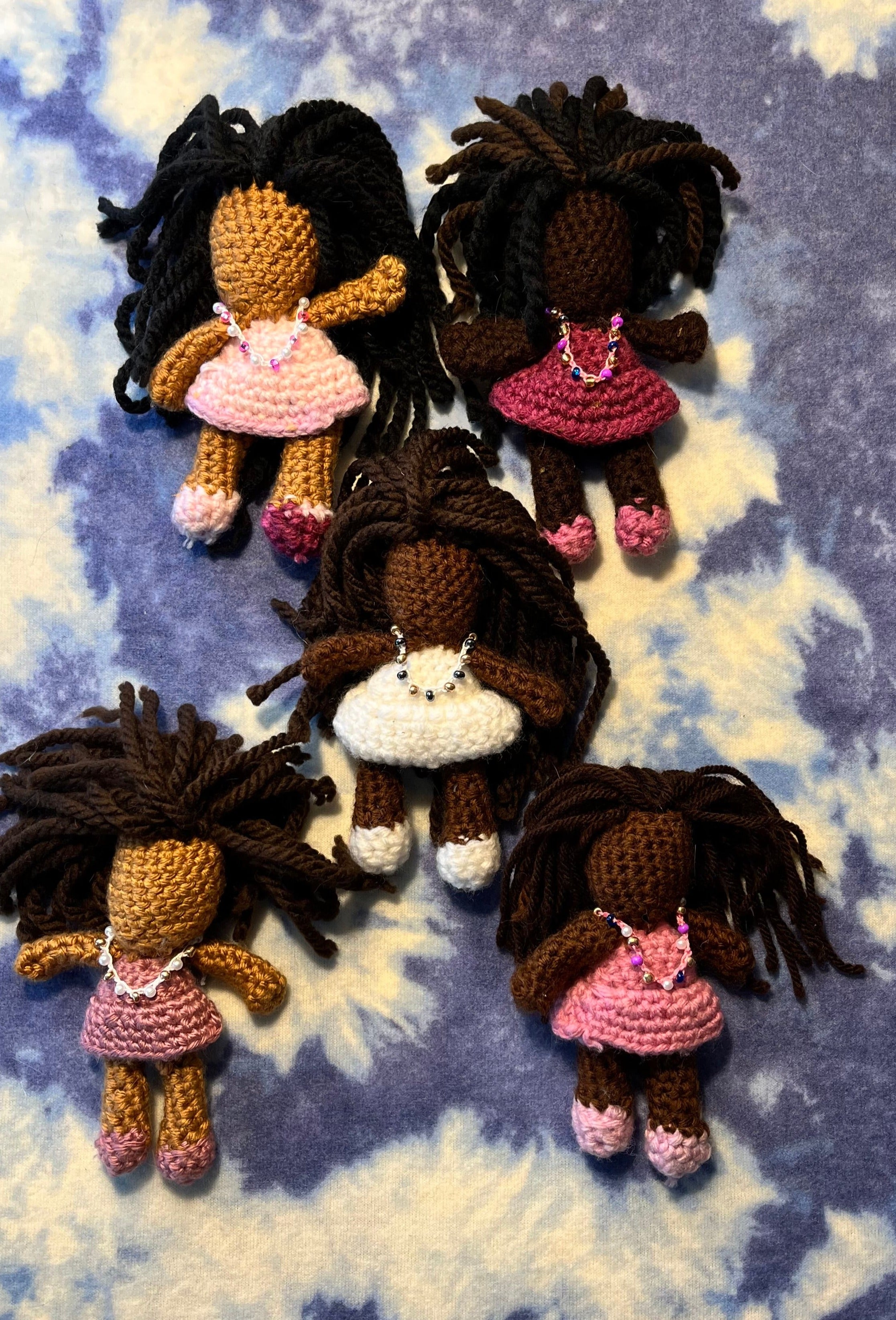 These.small crochet dolls have hand-tied black or brown yarn hair. They are wearing pink, mauve, or white dresses with matching shoes. 
