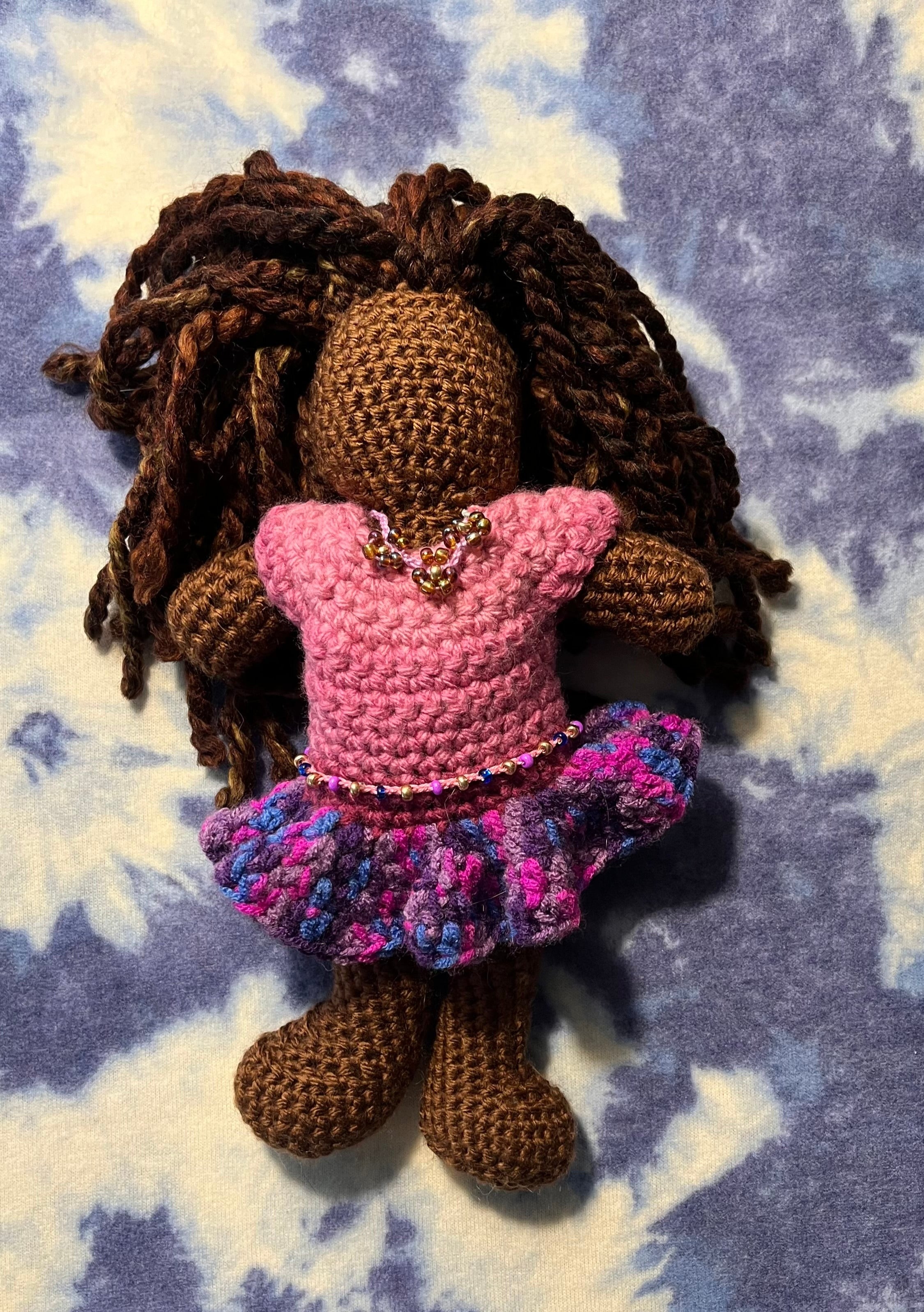 brown skinned crochet doll with brown twisted yarn hair. The doll is wearing a pink snd purple dress with a beaded necklace and waistbeads