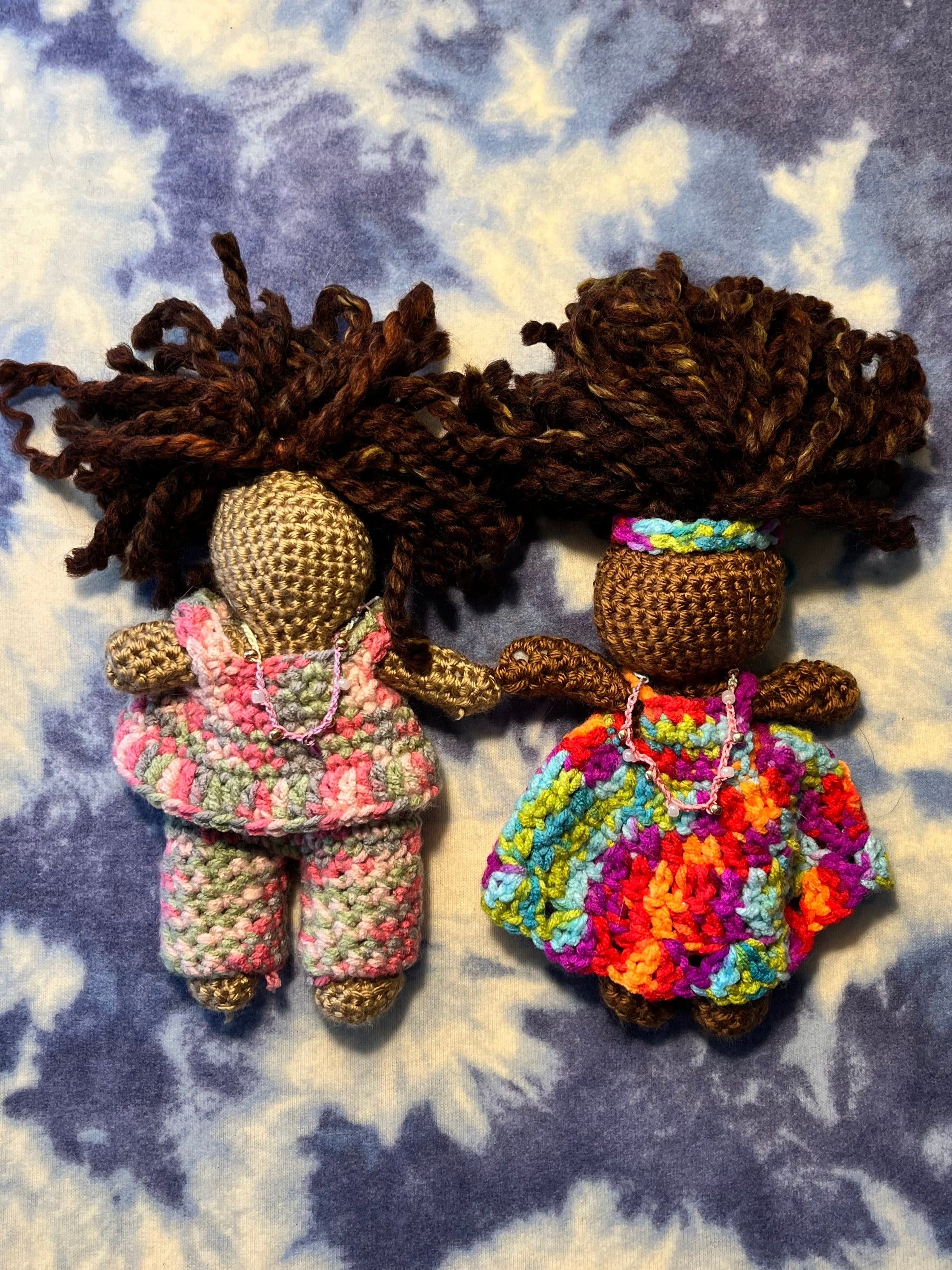 handmade crochet dolls with brown yarn hair. Doll on the left is tan skinned and is wearing a pink and green colored pant suit. Doll on the right is dark skinned and is wearing a orange, purple, green and blue multicolored dress with a matching headband. Both dolls have handbeaded necklaces. 