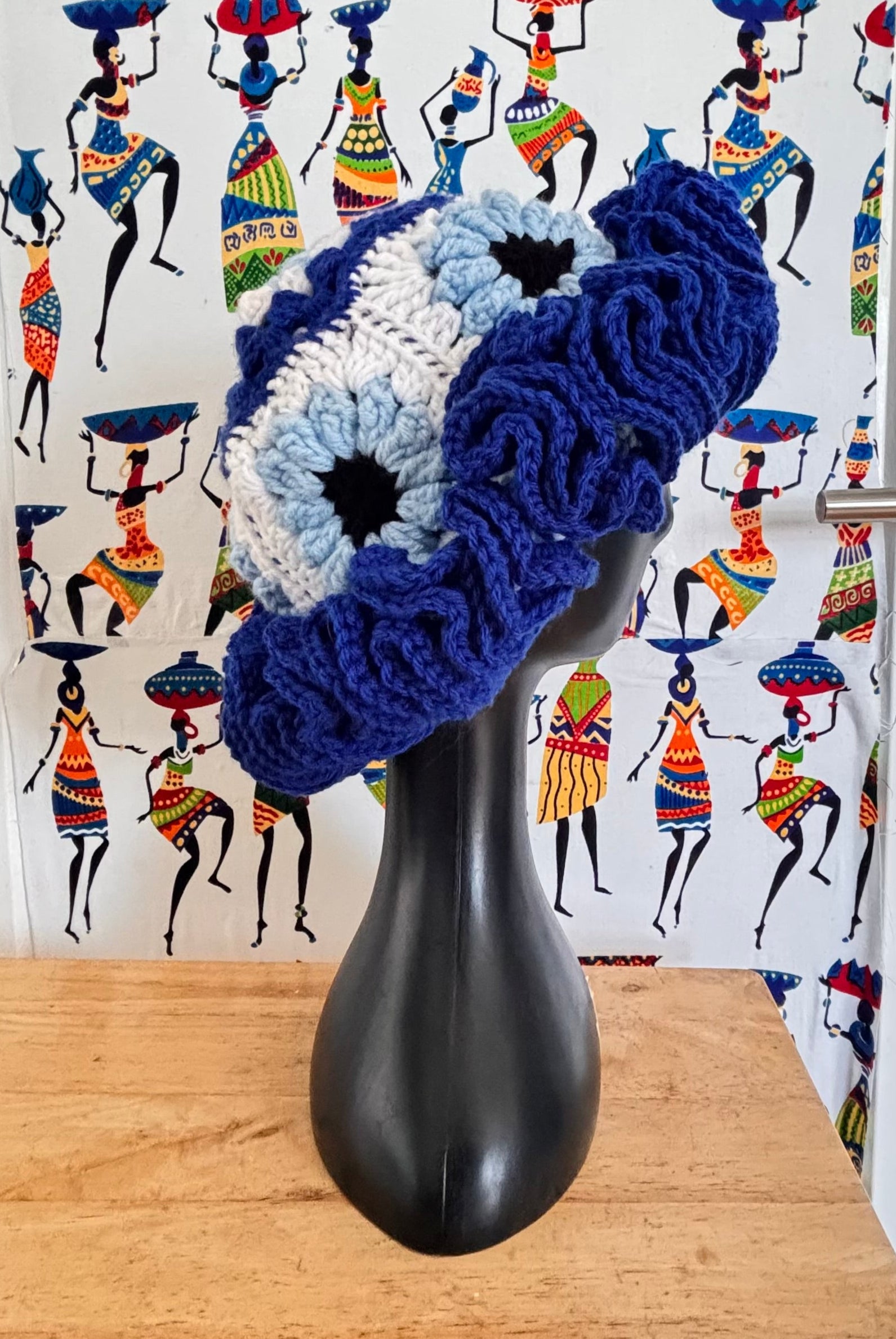 Decorative evil eye ruffle hat on a black stand with a colorful background featuring people in traditional attire.