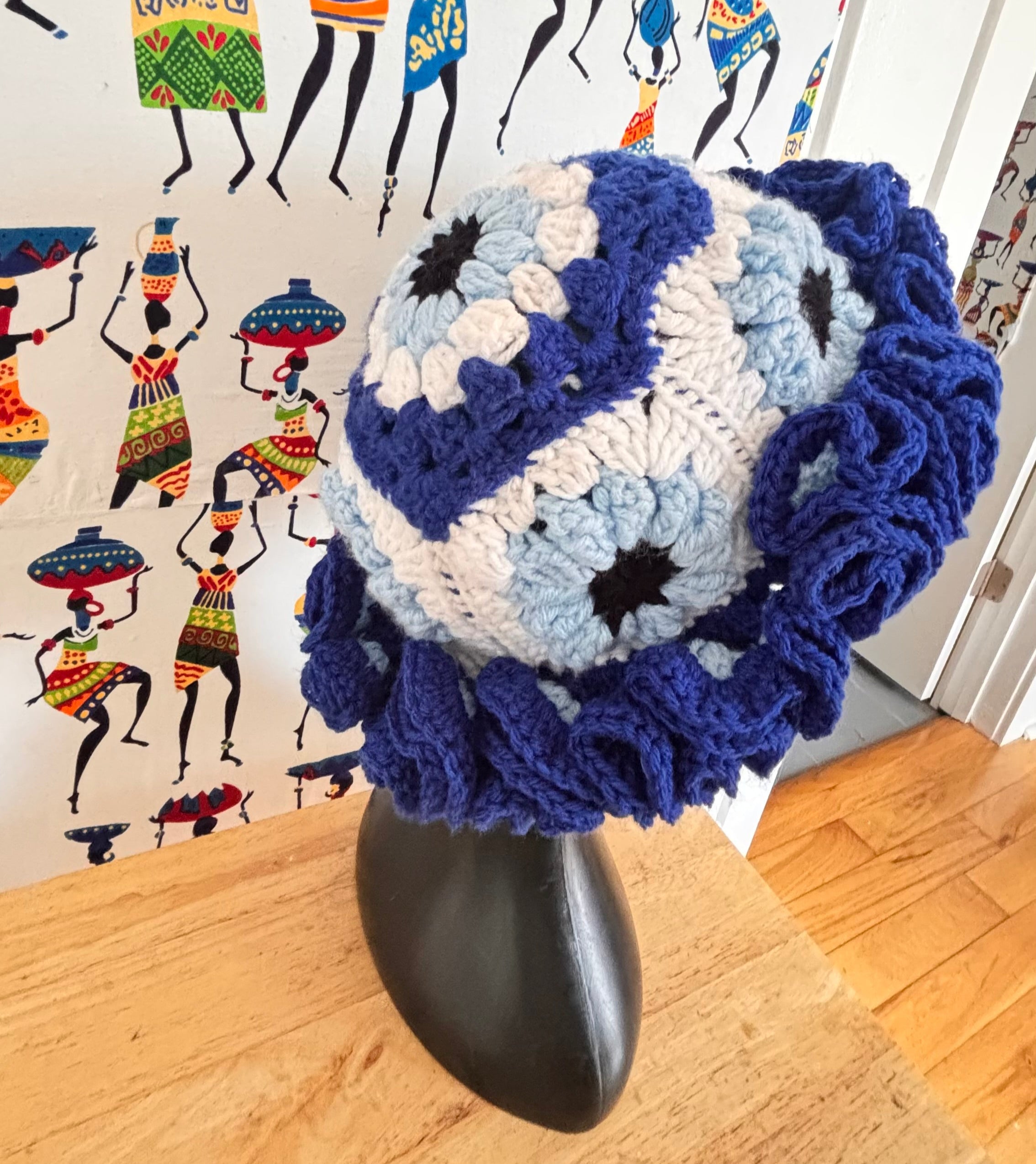 Crocheted blue and white evil eye granny square on a black stand with a colorful wall mural in the background